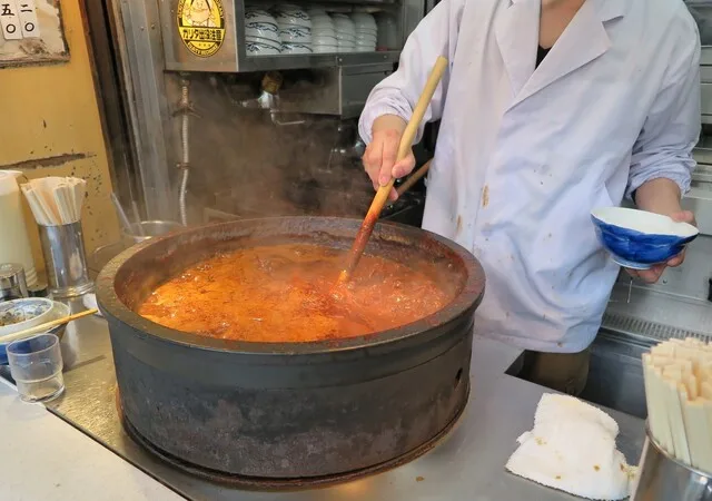 店先の寸胴で赤味噌のホルモン煮込みを混ぜる白衣の料理人
